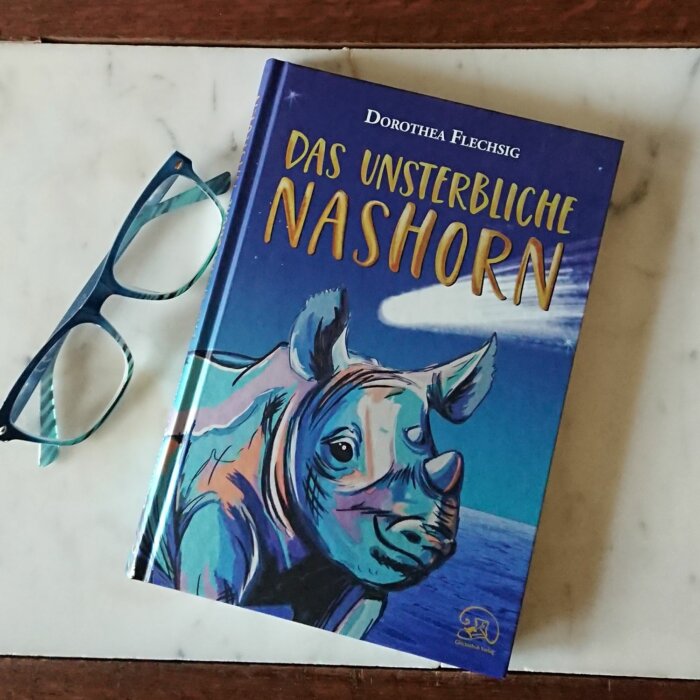 Buchcover: Das unsterbliche NAshorn. Phantastischer Kinderroman für Mädchen und Jungen ab 10 Jahre.
Das Bild zeigt das Buch auf hellem Untergrund, daneben eine farblich passende Lesebrille
