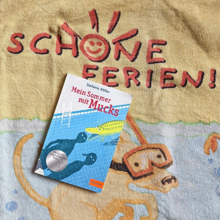 Taschenbuch Mein Sommer mit Mucks. Buch liegt auf einem Strandhandtuch mit der Aufschrift "Schöne Ferien"